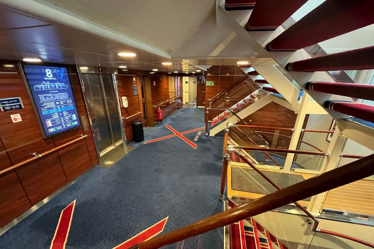 A stairwell and deck landing inside a Fjord Line ship, with wood panelling, carpeted stairs and a deck plan on the wall