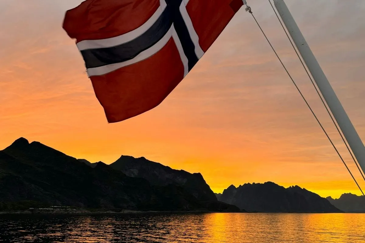 Det norske flag ved agterenden af et skib ved solnedgang, med fjelde og roligt vand bagved